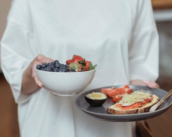 Person holding a bowl of fresh berries and a plate with avocado toast, displaying a healthy breakfast spread.